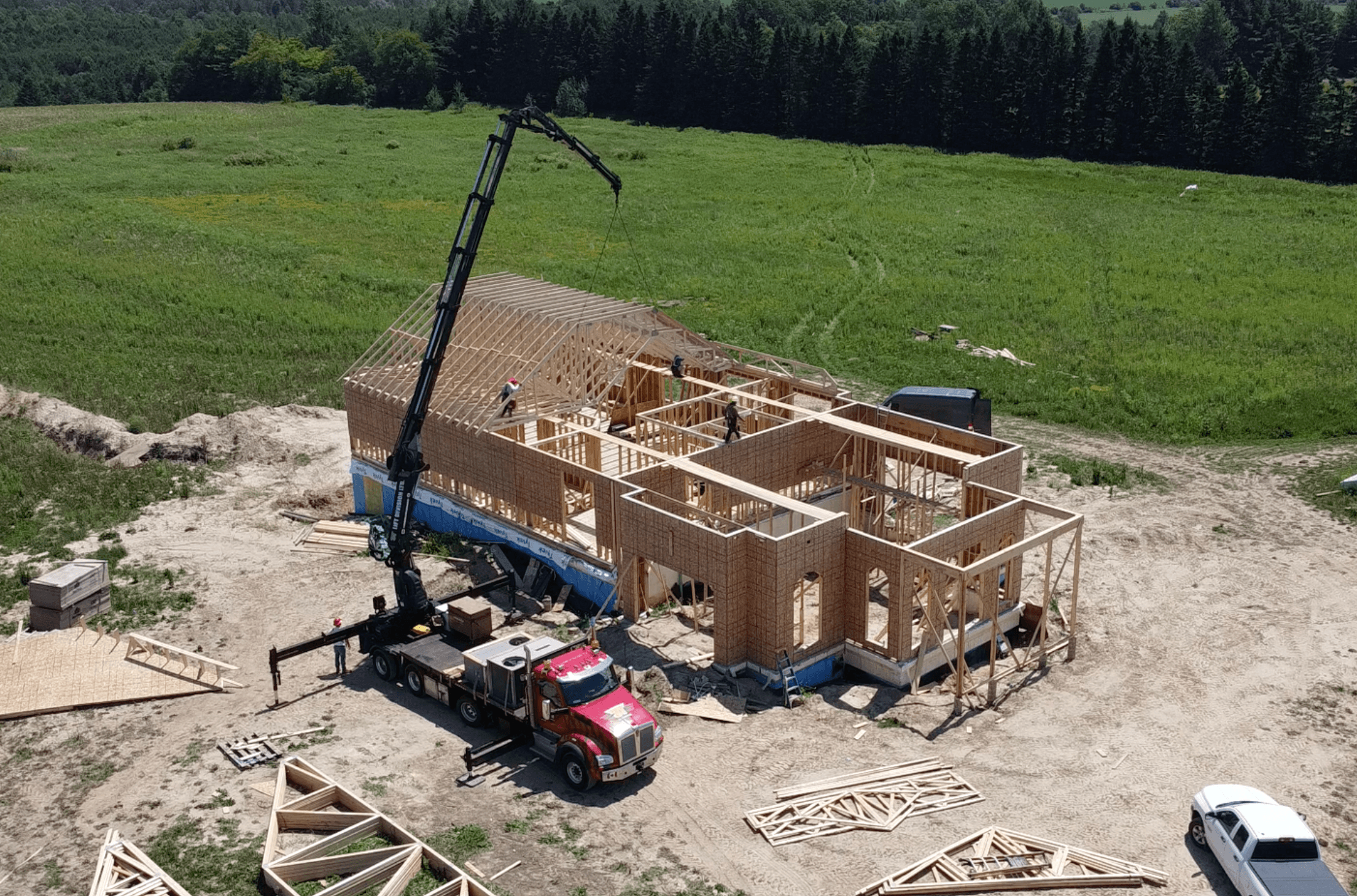 Aerial view of a red crane truck lifting roof trusses onto a wooden house frame.